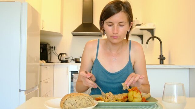 Woman sits at kitchen table eating stuffed paprika, enjoying homemade Balkan cuisine, turning meal into cultural moment shared in quiet domestic space.