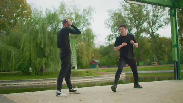 caucasian men practicing outdoor kick and footwork by lakeside, playful rivalry turns technical with roundhouse kick, evasive steps, drill and choreography under green structure, teammate challenger