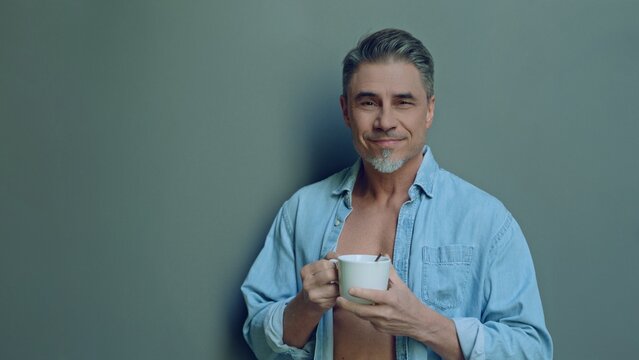Confident smiling mature man holding a coffee mug, standing in front of a gray wall, looking at camera. Mid adult male in 50s, open denim shirt, bearded, grey hair, friendly and approachable smile.