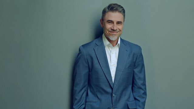 Well-dressed man in 50s standing against neutral backdrop, confidence and approachability with warm, charming smile. Business portrait of happy, friendly mature businessman smiling.