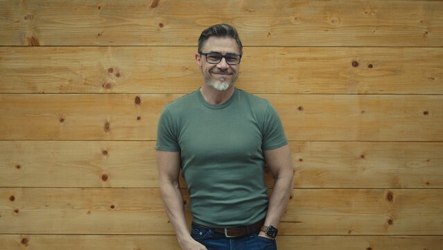 Portrait of happy mature man with eyeglasses and casual clothes standing, leaning against wooden wall, smiling. Mid adul, middle aged male in 50s. Copy space.