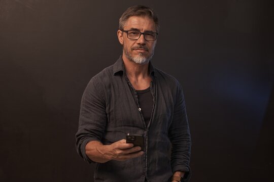 Portrait of mid adult man standing against dark background in gray casual clothes. Studio shot of middle aged male in glasses using mobile phone.