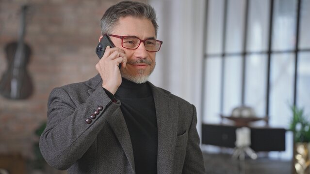 Smiling middle aged businessman talking on phone call at home. Mid adult older professional man in glasses communicating, using mobile. Telecommunication, business, manager skill.