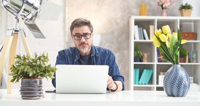 Bearded man working online with laptop computer at home sitting at desk. Home office, browsing internet, study room. Portrait of mature age, middle age, mid adult man in 50s.