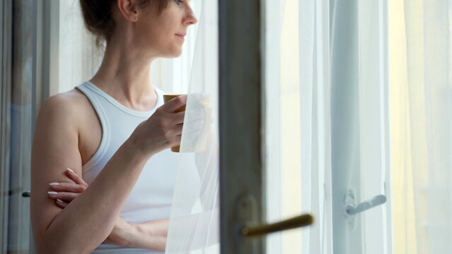 Young woman standing in window at home daydreaming and drinking morning coffee. Unrecognizable person.