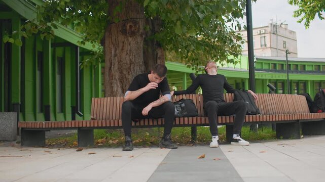 two white men sitting bench tense urban scene college student and delivery rider exchange awkward silence then quiet support wide park shot with large tree and green pavilion backpacks on bench,