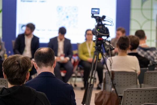 Business panel recorded during seminar, audience watches while camera captures speakers, professional conference atmosphere.