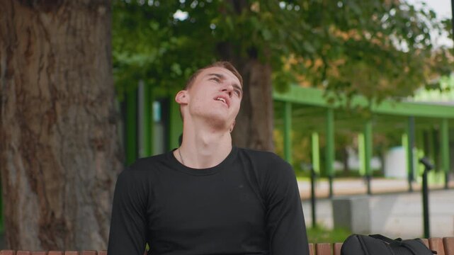 white male gazing upward on bench near green pavilion and skyline, subtle neck stretch and thoughtful expression, solo backpack beside him, soft overcast light, calm reflective mood