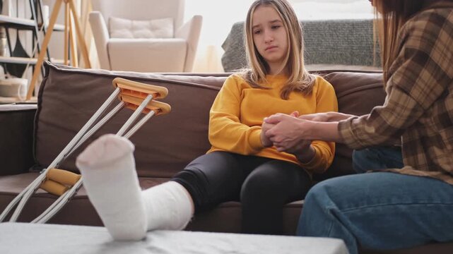 Girl with broken leg in cast sitting with mother on couch at home. Sad teenage kid recovering from broken leg. Girl using cast crutches for quick rehabilitation. Mom supporting daughter