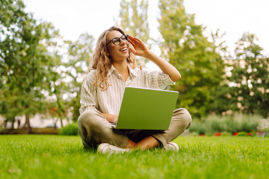A happy woman sits on the grass in a park with a laptop on a sunny day. A female freelancer works on a laptop in a park at sunset. Concepts for remote work, education, or leisure.