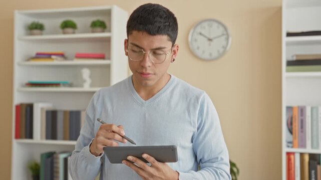 Man holding tablet and stylus, writing on touchscreen and glancing upward in a study, wearing glasses and sweater with books and clock visible; focused concentration.