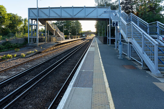 Elevated view of a quiet train platform with parallel tracks, a small station building, and lush trees. A calm, everyday transit scene capturing travel,