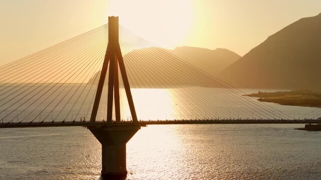 Famous Rio-Antirrio bridge silhouetted against a golden sunset, with shimmering water reflections and distant mountains creating a breathtaking and scenic hellenic landscape