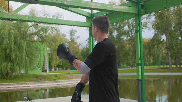 white male posing with gloves lakeside near green pavilion, tattooed forearm and calm expression, pond reflections and afternoon sunlight, cooldown walk and quiet confidence, urban park contrast