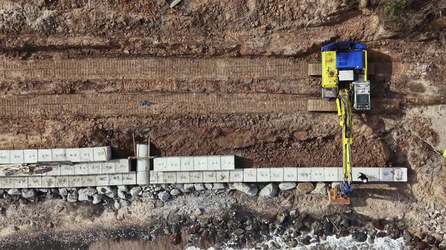 Heavy machinery works on a coastal project. Workers place large concrete blocks along the shore. The setting features an exposed area near the water with rocks scattered on the ground.