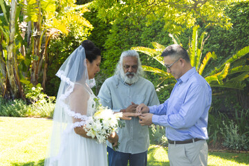Couple with Indian officiant exchanging rings on sunlit lawn, bride holding white bouquet and veil © wavebreak3