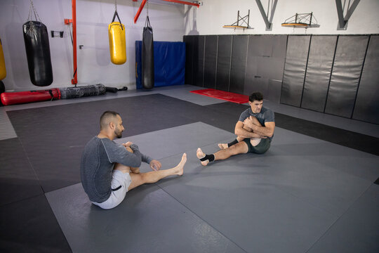 Diverse male training partners stretching on mats in gym wearing athletic shorts with ankle tape