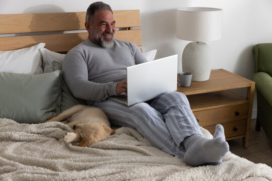 Senior man sitting on bed typing on laptop in pajama pants with sleeping dog and mug