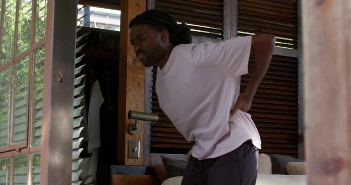 African American man sitting on bed pressing lower back in pain, standing at doorway rubbing hip