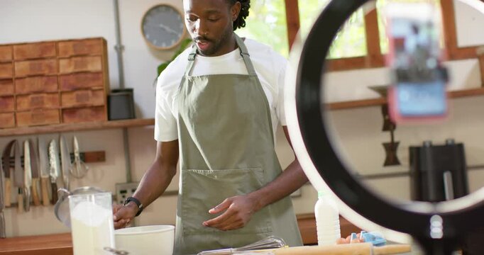 African man in apron filming on phone, sieving flour, cracking egg in kitchen for lesson