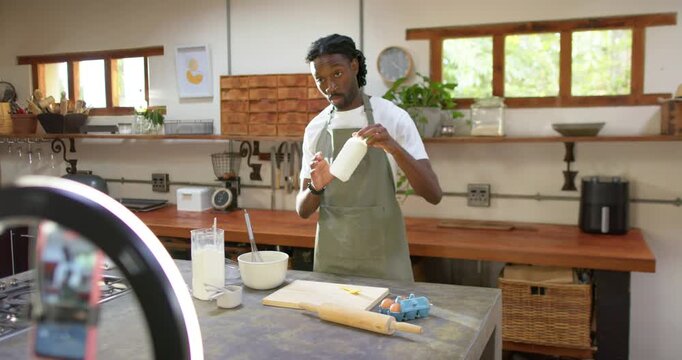 Adult African American man whisking at kitchen island with ring light filming recipe, pouring milk