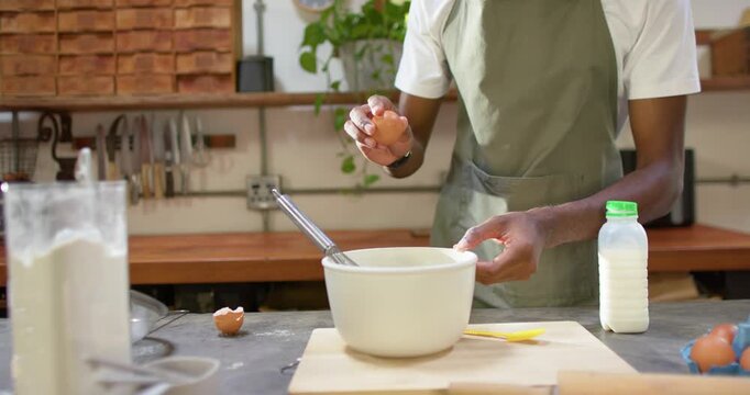 African man in green apron preparing batter by cracking eggs into mixing bowl at kitchen counter