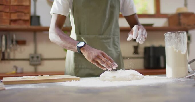 African American man in apron kneading dough at counter reaching flour jar, shaping loaf for baking