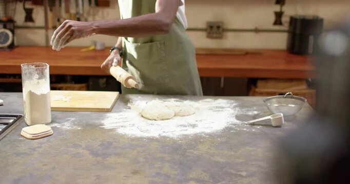 African American man in apron spotting dough, dusting flour from jar, tapping rolling pin to roll