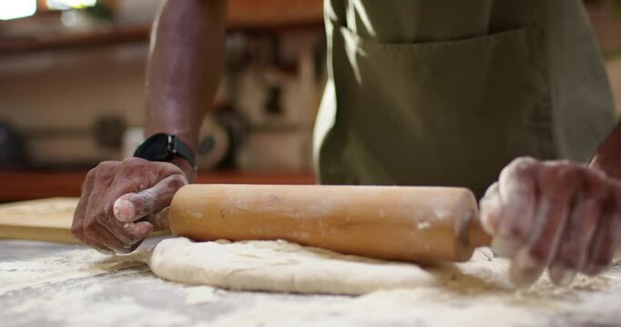 Adult African man in green apron on floured counter placing rolling pin flattening dough for baking