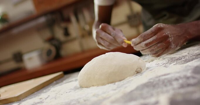 Male baker noticing proofed dough, scoring loaf with yellow tool on floured wood counter for baking