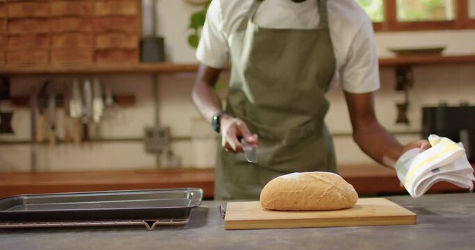 African American man wearing apron placing loaf on board at counter slicing with knife to bake