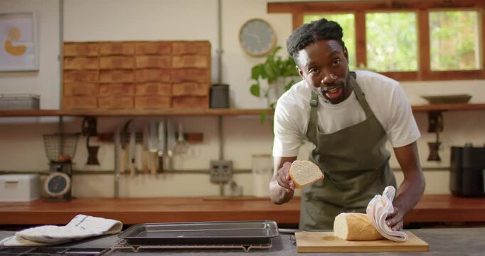 Man in olive-green apron lifting bread slice from loaf addressing camera while placing on tray