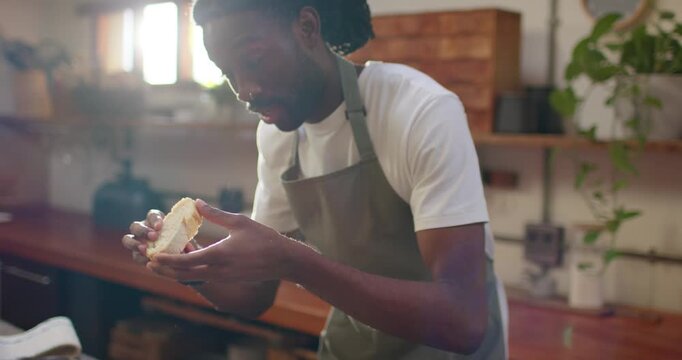 Adult man wearing olive apron holding halved roll, inspecting, smelling and tasting on wood counter