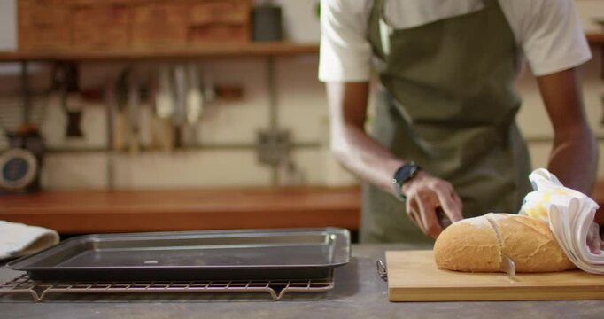 Adult African man green-aproned slicing loaf on counter with serrated-knife steadying towel to bake