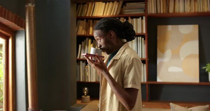 Man in beige shirt glancing at window, holding cup on saucer, raising mug smelling beverage