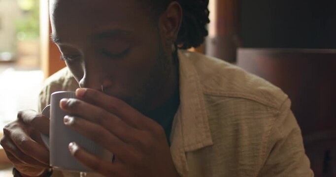 African American man holding mug lifting inhaling sipping at cafe booth by window in light shirt