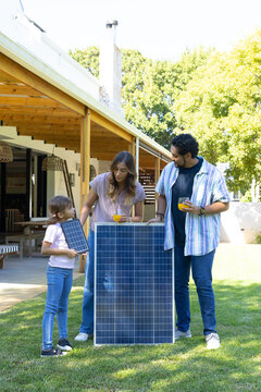 Diverse family standing on backyard lawn by large solar panel, child holding small panel