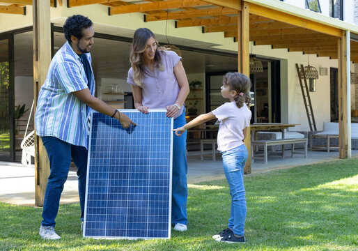 Family with parents and daughter holding upright solar panel, pointing and talking on lawn