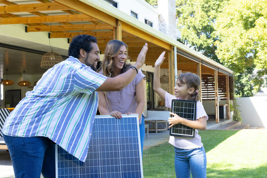 Family of three standing on lawn holding solar panels by wooden patio and pergola