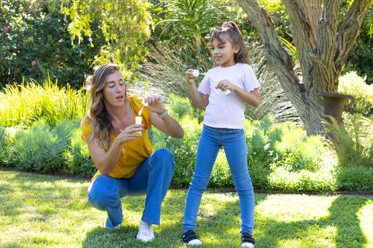 Mother and daughter kneeling blowing soap bubbles with bubble bottles and wands on sunlit lawn