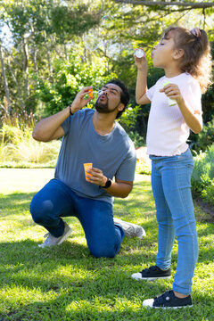 African American father and daughter blowing soap bubbles on grassy lawn, holding bubble containers