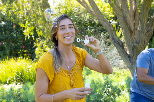Adult couple standing in garden, woman in mustard blouse holding bubble wand, creating bubbles