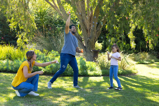 Diverse family playing with bubble wands and soap bubbles on grassy lawn under large shade tree