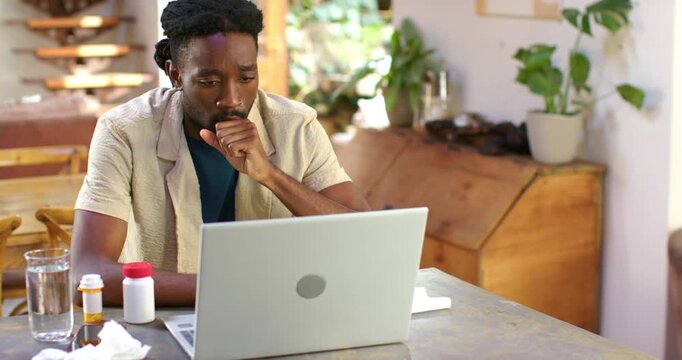 Man coughing while feeling unwell, monitoring laptop for work at table with pill bottles
