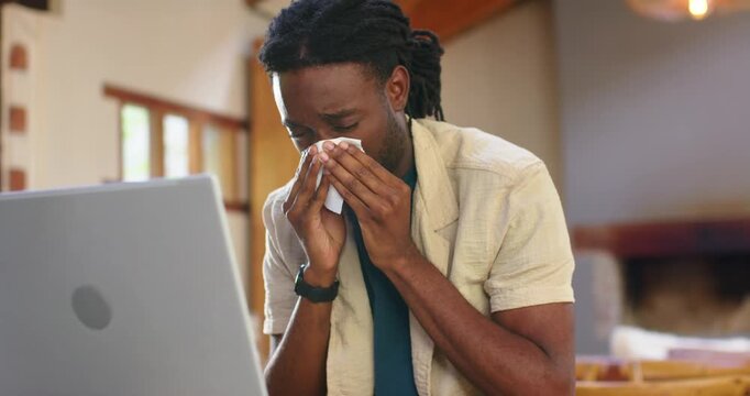 Man sitting at table at home, glancing at laptop, blowing nose with tissue, refocusing on work