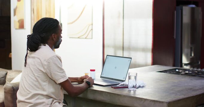 Man reacting to laptop, reaching for pill bottle with red cap, checking dosage at kitchen island