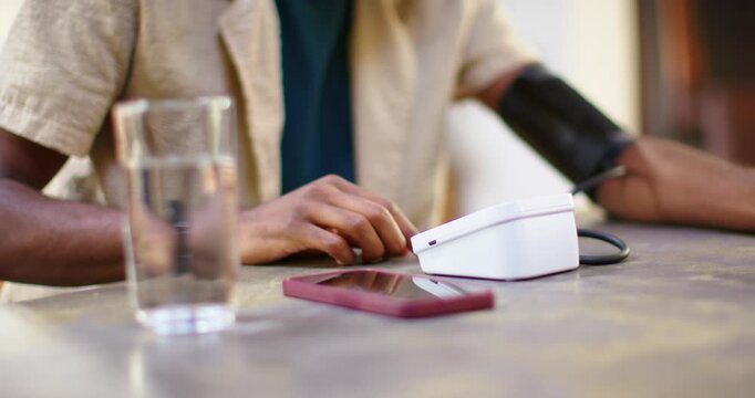 African American man pressing BP button to check BP, adjusting cuff, drinking water at table
