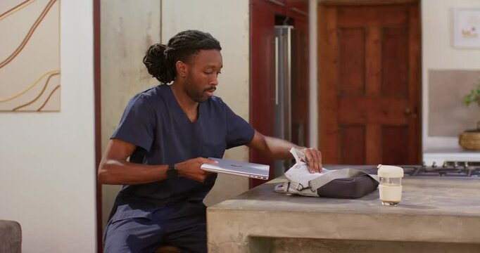 Adult African man in scrubs at counter reaching into gray backpack, removing silver laptop to work