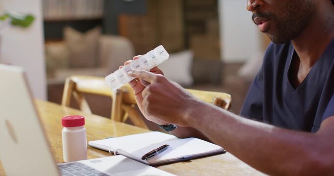 Seeing rx bottle adult African man in scrubs at table pointing at weekly pillbox confirming dose