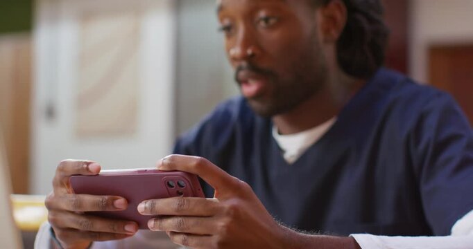African American man sitting at home wearing navy scrubs, replying on phone and glancing at laptop